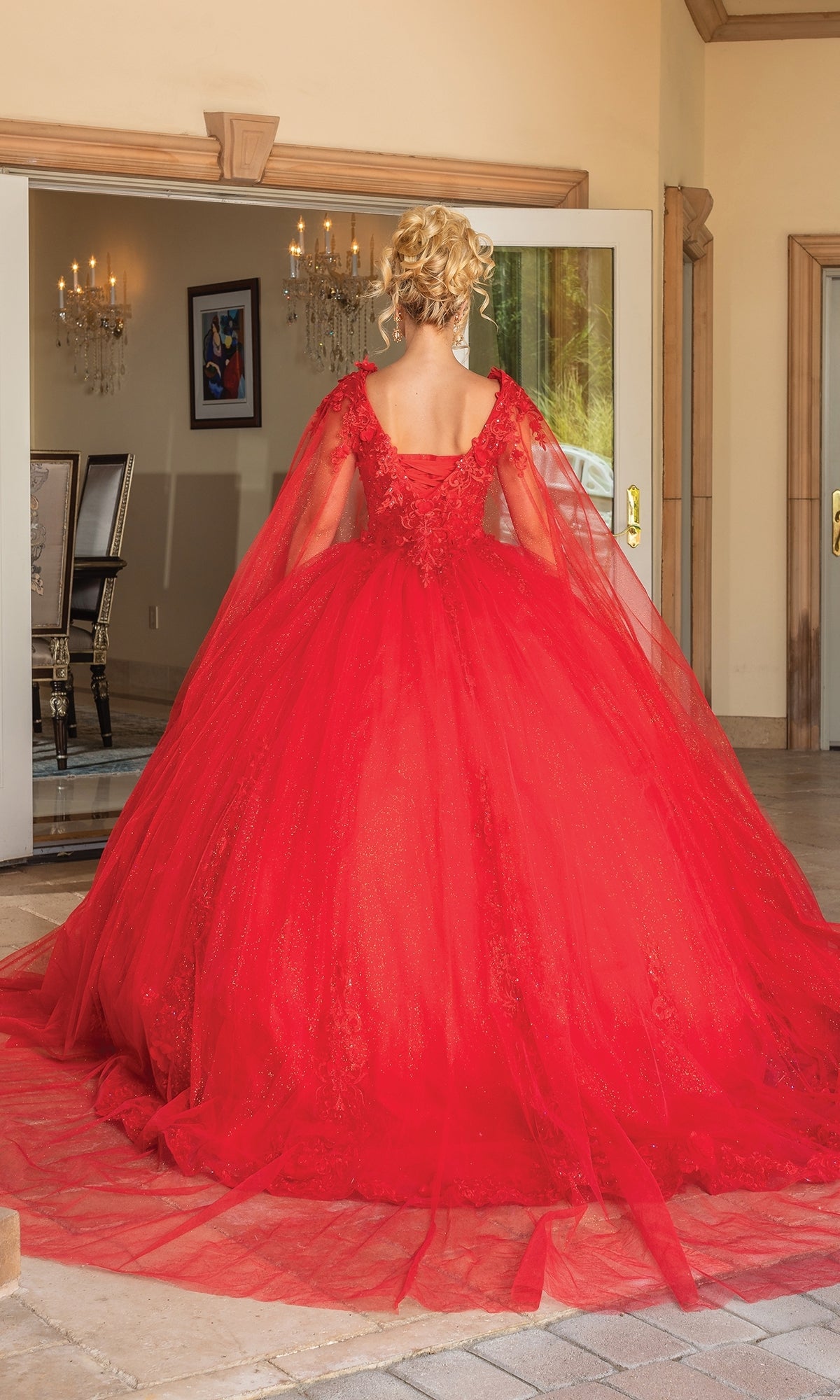 A woman with blonde hair in an updo wears the Quinceanera Dress 1678 by Dancing Queen, a voluminous red ball gown with lace and tulle, standing indoors facing away from the camera.